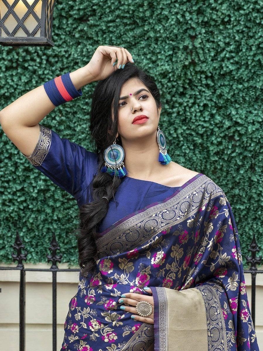 Woman in navy blue floral saree with gold border, styled with statement earrings and bangles, against green hedge backdrop