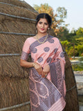 Woman in pink printed saree with floral patterns, standing by thatched hut, elegant ethnic wear
